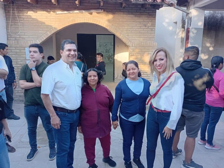 Hugo Adalberto Velázquez Moreno sonriente en camisa blanca, acompañado de dos mujeres. Ambiente urbano con paredes de ladrillo expuesto.
