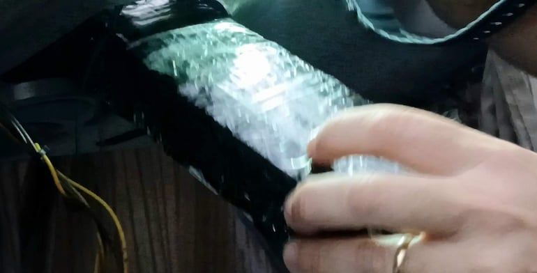 Una mano sostiene un objeto parcialmente envuelto en plástico en un ambiente oscuro con cables visibles.