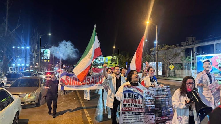 Grupo de médicos con batas marchando bajo luces nocturnas, algunos levantan carteles y voces de protesta en la calle.