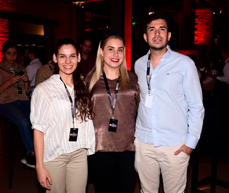 Una mujer con blusa blanca sonríe junto a otra con blusa marrón y un hombre en camisa celeste, en un bar con luces tenue.