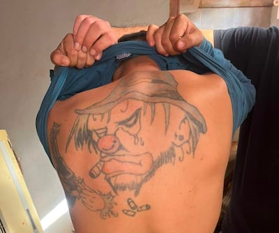 Hombre brasileño levantando su camiseta para mostrar un tatuaje de payaso triste, con lágrimas y un cigarro, en un ambiente iluminado.