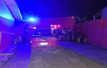 Ambiente nocturno con vehículo rojo en primer plano, luces azules y un grupo de personas en un callejón.