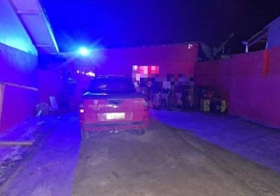 Ambiente nocturno con vehículo rojo en primer plano, luces azules y un grupo de personas en un callejón.