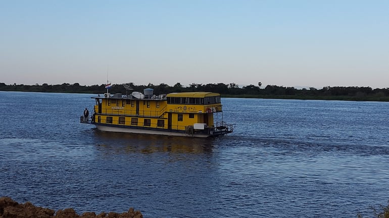 Barco hotel de color amarillo en un río rodeado de árboles, bajo un cielo despejado.