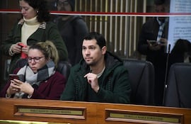 Pablo Callizo con abrigo verde oscuro y micrófono, Paulina Serrano con gafas y abrigo marrón, en sala oficial atenta a la discusión.