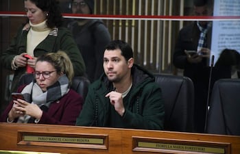 Pablo Callizo con abrigo verde oscuro y micrófono, Paulina Serrano con gafas y abrigo marrón, en sala oficial atenta a la discusión.