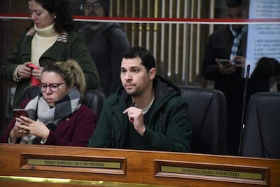 Pablo Callizo con abrigo verde oscuro y micrófono, Paulina Serrano con gafas y abrigo marrón, en sala oficial atenta a la discusión.