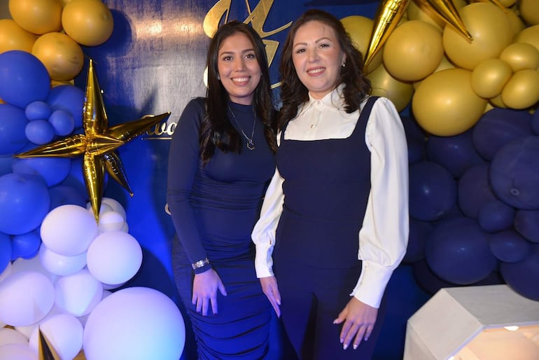 Dos mujeres sonrientes, una en vestido azul oscuro y otra en blusa blanca con overol azul, en un ambiente decorado con globos.