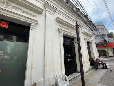 Tres personas sentadas en sillas blancas y metálicas, en una esquina de un edificio blanco, con un ambiente urbano desolador.