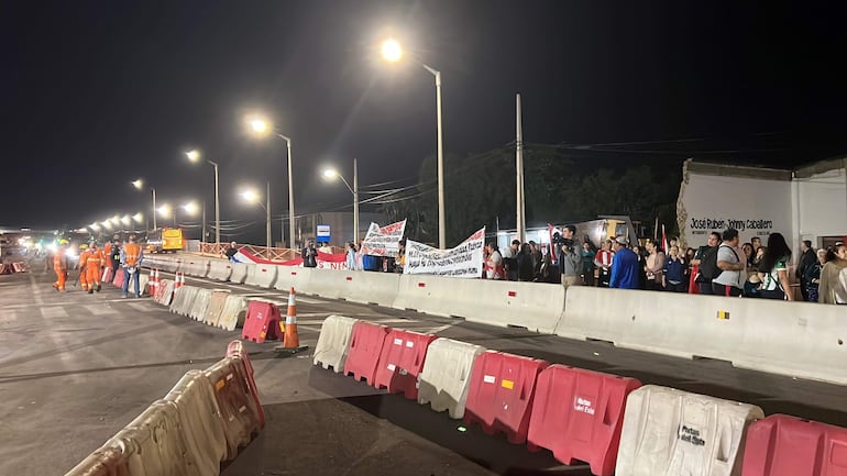 Protesta nocturna con grupo de personas y trabajadores de seguridad en ropa naranja, sosteniendo pancartas blancas con texto negro.