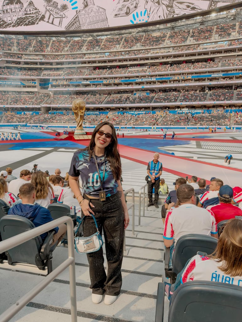 Mujer con camiseta número 10 y bolso sonríe en estadio lleno, con trofeo y pantalla de fondo.