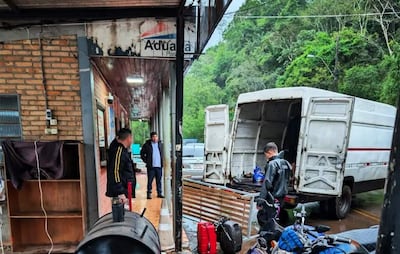 Tres hombres en ambiente lluvioso: uno de pie con chaqueta oscura, otro con abrigo claro y un joven en chaqueta de trabajo inspeccionando un vehículo.
