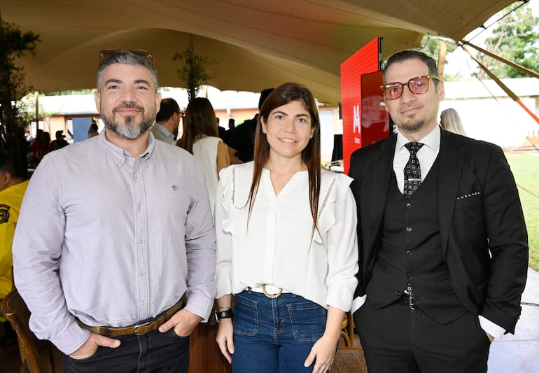 HOMBRE  sonríe vestido con traje oscuro mientras una mujer de blusa blanca lo observa, en evento bajo carpa con otras personas al fondo.