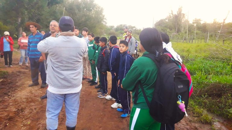 Grupo de alumnos y adultos en campo abierto, algunos niños en uniforme deportivo escuchando a un hombre de espaldas conversando con ellos.