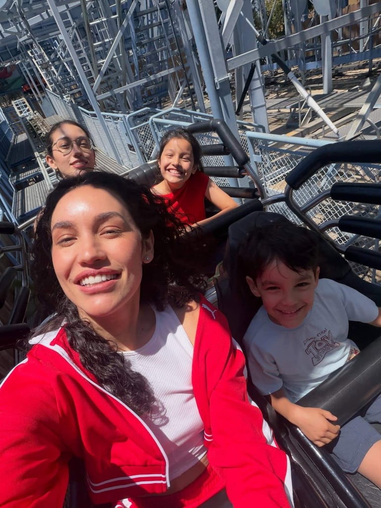 Mujer con chaqueta roja y niños sonrientes en montaña rusa, rodeados de estructuras metálicas del parque.
