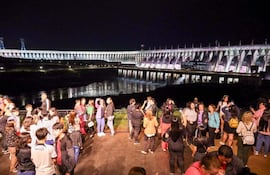 Grupo de personas conversando y tomando fotos, con vestimenta casual, frente a la iluminada represa de Itaipú.