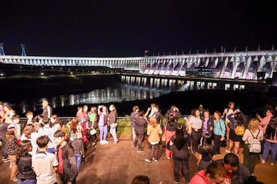 Grupo de personas conversando y tomando fotos, con vestimenta casual, frente a la iluminada represa de Itaipú.
