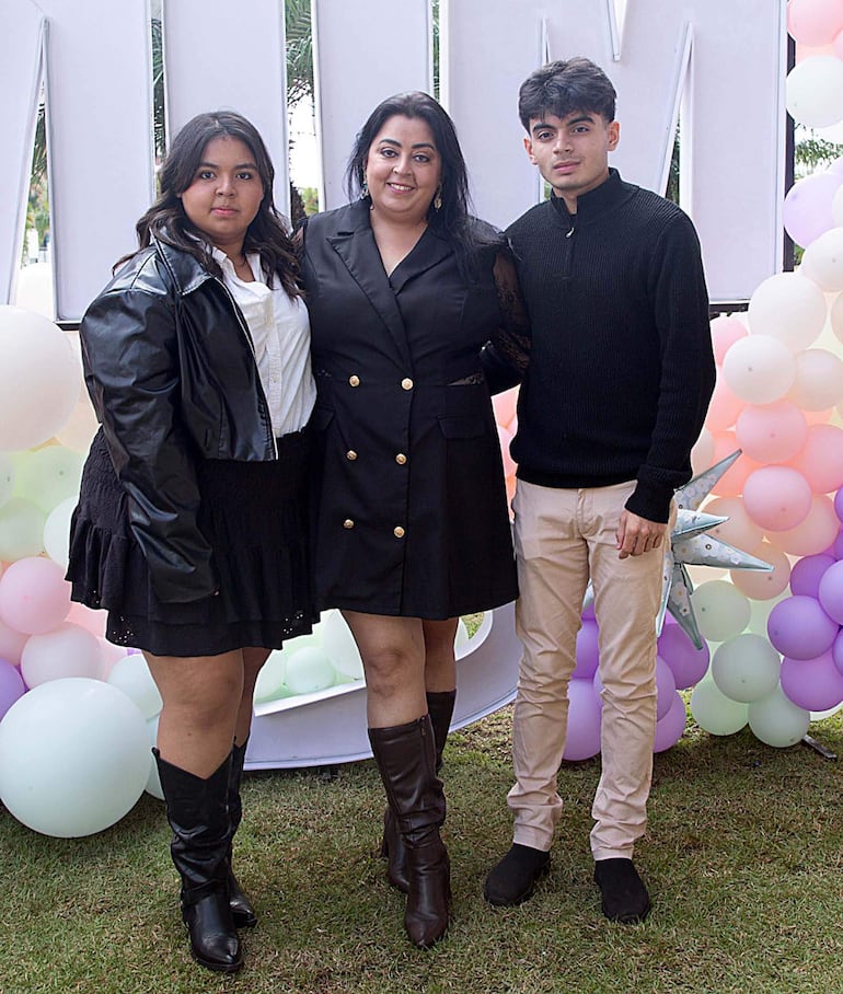 Joven con chaqueta negra y falda, mujer con chaqueta de botones dorados y joven en suéter negro sonríen ante fondo de globos.