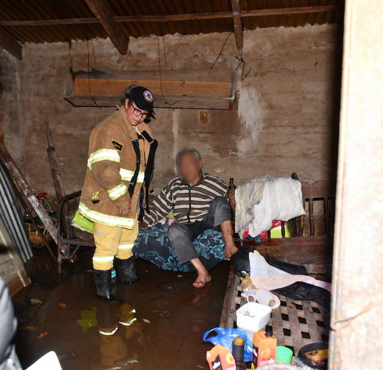 Bombero de pie en uniforme asiste a hombre mayor sentado en sofá, con ambiente interior dañado por agua.