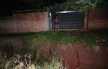 Mujer vestida con abrigo y pantalones cortos, al borde de un precipicio, rodeada de vegetación escasa y barro en un ambiente nocturno.