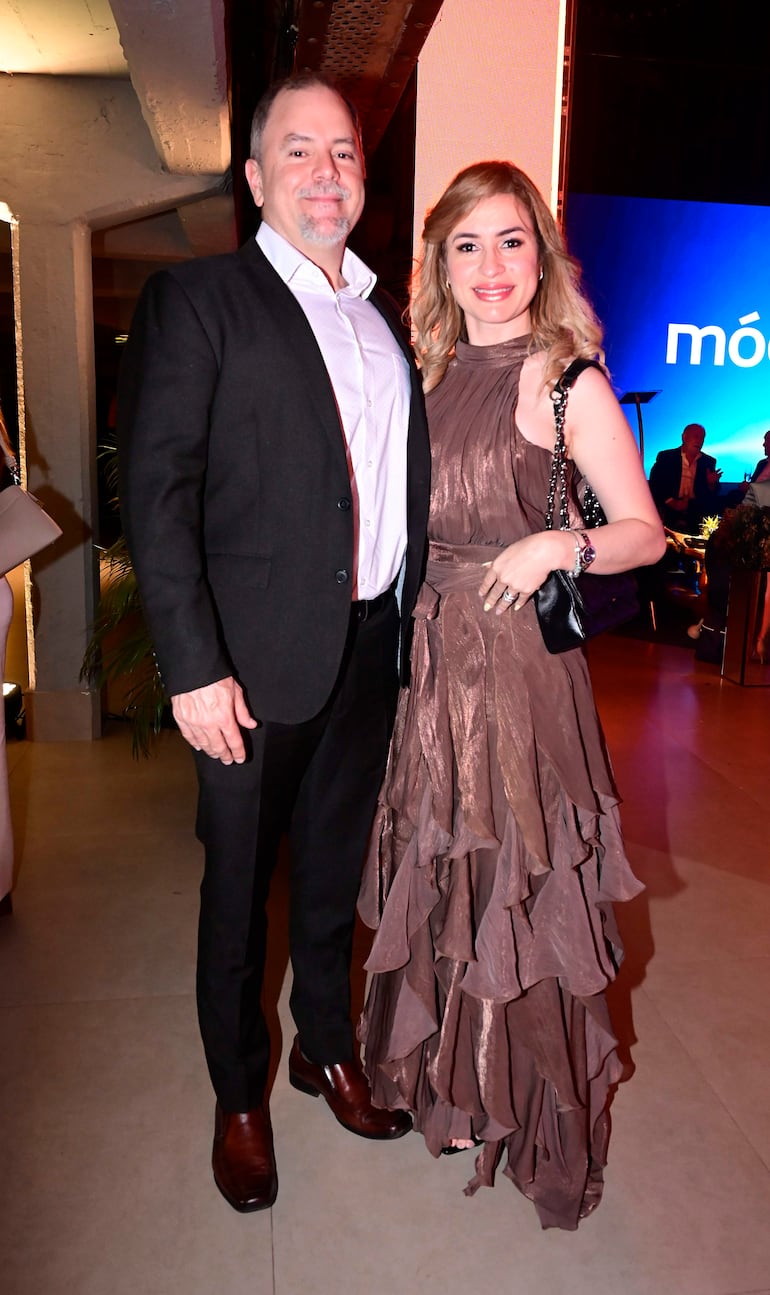 Hombre en saco oscuro y mujer en vestido marrón elegante sonríen en evento iluminado con pantalla azul de fondo.