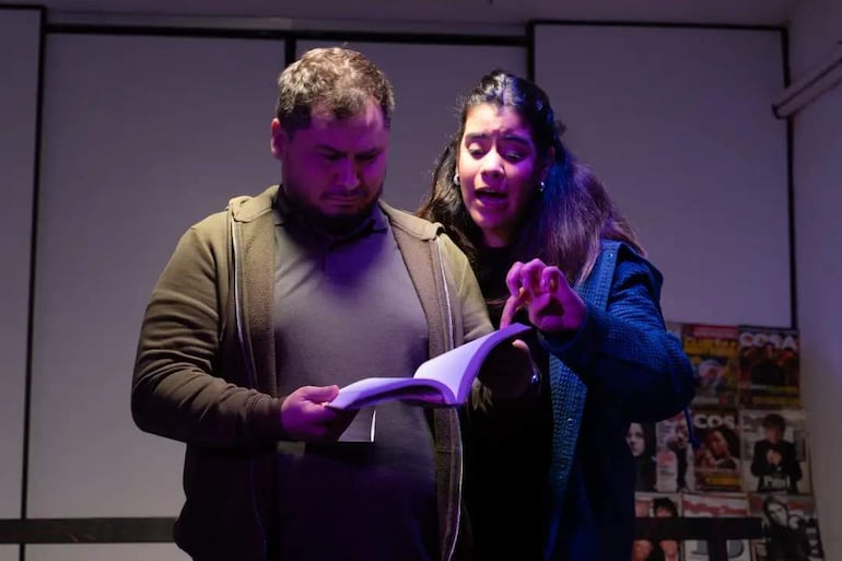 Mujer de pie señalando un libro, hombre sentado con expresión confusa, ambiente cálido y revistas en la pared.