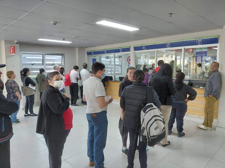 Mujer sonriente en primera fila, personas con mascarillas esperando atención en mostradores de farmacia.