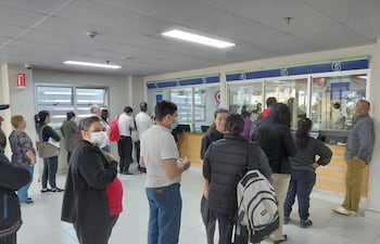 Mujer sonriente en primera fila, personas con mascarillas esperando atención en mostradores de farmacia.