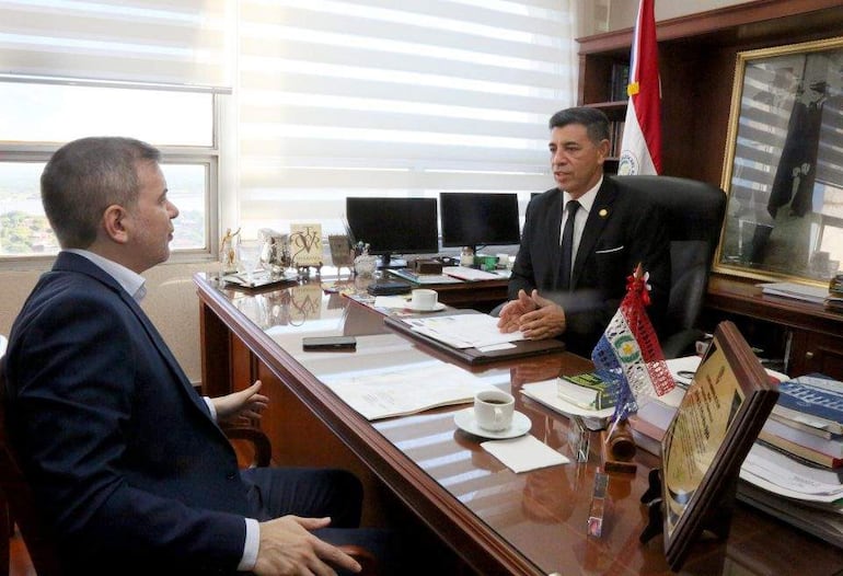 Víctor Ríos Ojeda en traje oscuro escucha atentamente a José Duarte, quien gesticula mientras habla, rodeados de documentos y una taza de café.