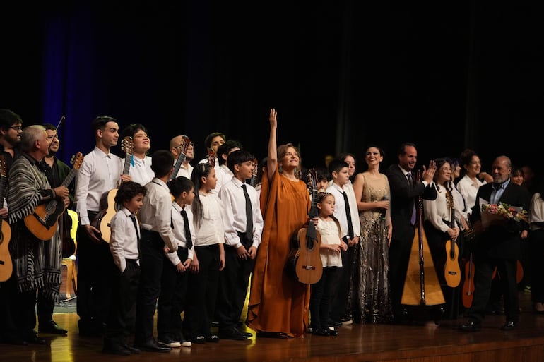 Berta Rojas en túnica naranja levantando un brazo, rodeada de jóvenes músicos sonriendo y aplaudiendo en un escenario iluminado.