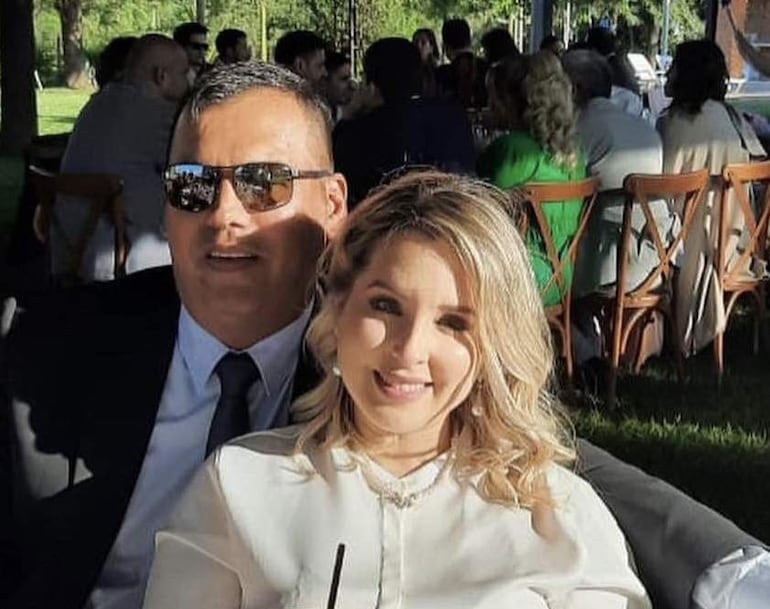 Pedro Alejandro Díaz Verón con traje oscuro y gafas de sol, junto a una mujer de cabello rubio y blusa blanca, sonríen en un ambiente festivo.