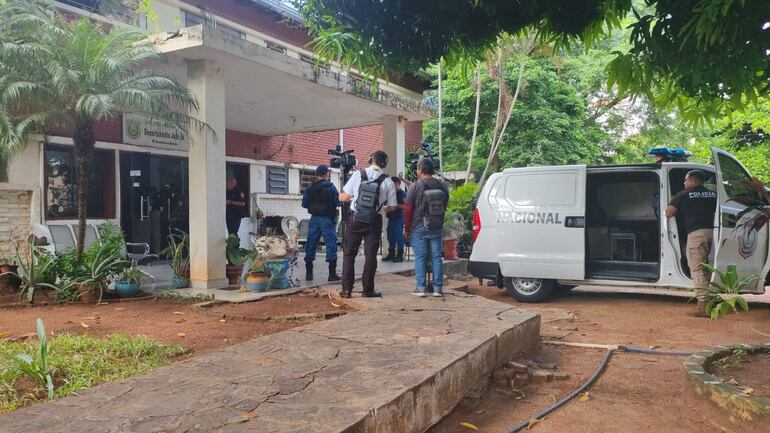 Hombre en camiseta blanca habla a un grupo de periodistas frente a un edificio de ladrillo; furgoneta policial abierta a la derecha.