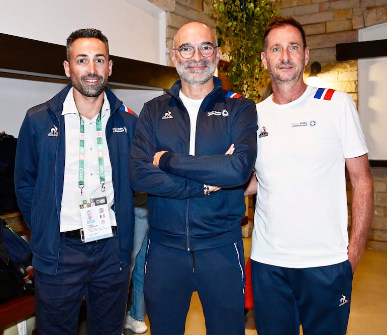 Hombres de pie en evento formal: uno con chaleco azul de la selección francesa, otro con gafas y expresión solemne, el tercero en camiseta blanca.