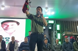 Eduardo Dejesus en pie, con micrófono y bandera, hablando mientras un grupo lo observa con atención en un ambiente decorado con luces verdes.