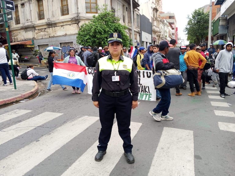 Oficial de policía en uniforme de pie, al centro, mirando hacia la cámara, mientras una mujer sostiene una bandera paraguaya.