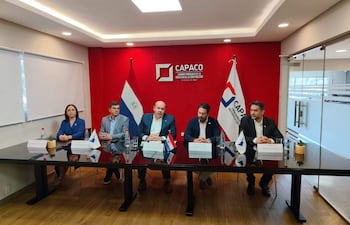 Reunión con cinco hombres y una mujer en mesa, con botellas de agua y plantas; fondo rojo y banderas presentes.