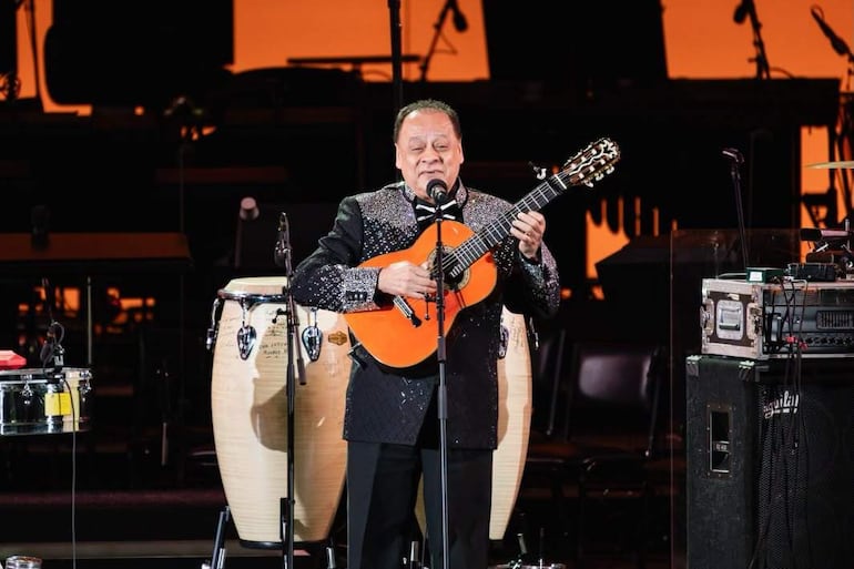 Gabriel Vargas en abrigo brillante, cantando y tocando guitarra, con congas y tambor en el escenario iluminado.