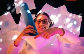 Mujer con gafas y dos coletas, sonriendo mientras sostiene un objeto azul y revisa dibujos en hojas de papel.