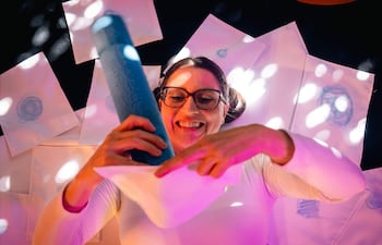 Mujer con gafas y dos coletas, sonriendo mientras sostiene un objeto azul y revisa dibujos en hojas de papel.