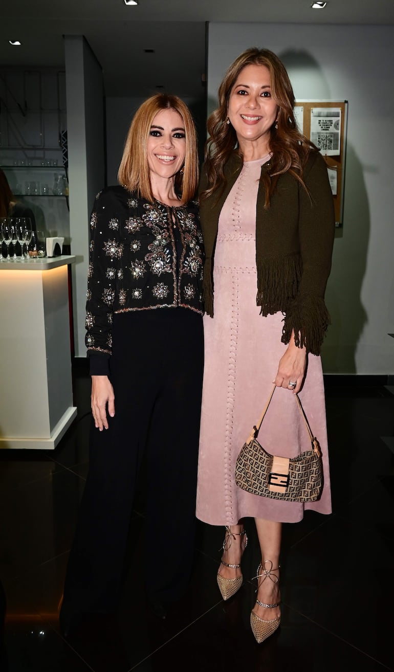 Dos mujeres sonrientes, una en blusa negra y otra en vestido rosa pálido, posan en un evento elegante con fondo de barra de bebidas.