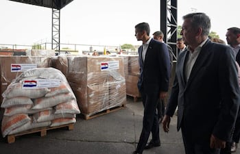 Dos hombres en traje oscuro, uno hablando y otro escuchando, rodeados de cajas y sacos de arroz en un ambiente interno.
