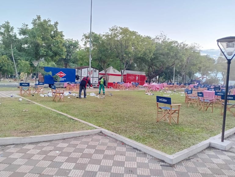 Dos personas, una con abrigo negro y otra con uniforme verde, recogen basura en la Plaza de Armas rodeada de árboles.