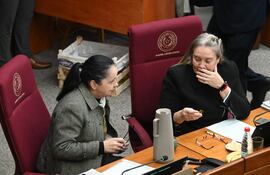 Gladys Lucía Mendoza de Esparza con abrigo claro discute, mientras Ramona Yolanda Paredes escucha atentamente en ambiente legislativo.