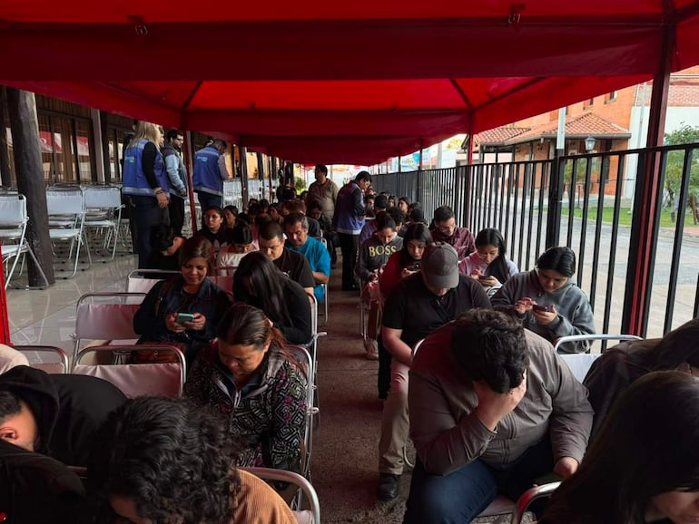 Grupo de personas sentadas bajo un toldo rojo, mirando sus teléfonos con expresiones de tristeza y tranquilidad en un ambiente exterior.