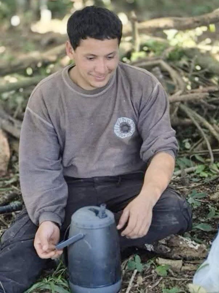 Joven con sudadera gris, sentado entre hojas verdes, sosteniendo un recipiente negro en un entorno natural.