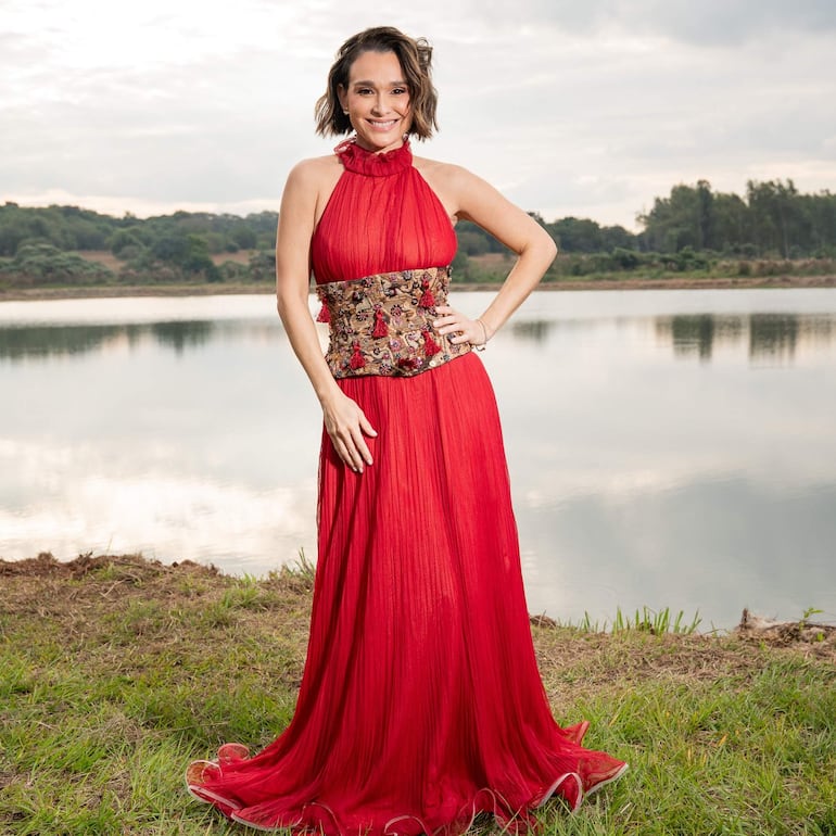 Emma Viedma sonríe en un elegante vestido rojo, posando junto a un lago con vegetación y colinas de fondo.