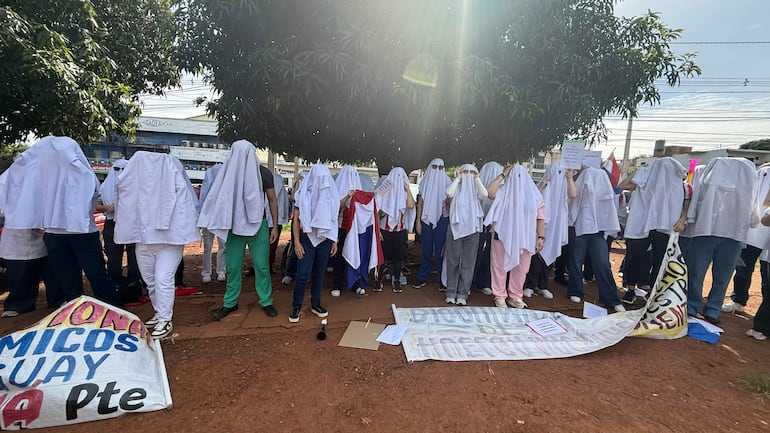 Veinte bioquímicos con batas blancas protestan en plaza, sosteniendo carteles y una pancarta enrollada en el suelo.