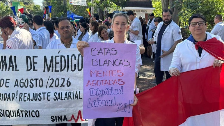 Mujeres y hombres en batas blancas sosteniendo pancartas en una protesta, con expresiones de determinación y un ambiente de manifestación.