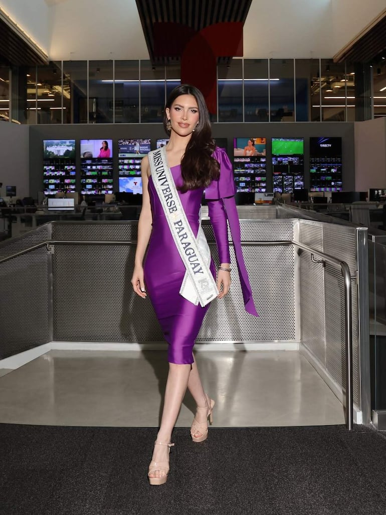 Gretha Matiauda sonríe mientras camina con vestido morado y banda de Miss Universo Paraguay en un entorno moderno.