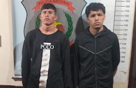 Dos hombres de pie con manillas de seguridad, uno vestido con camiseta negra y el otro con abrigo negro, junto al escudo policial.
